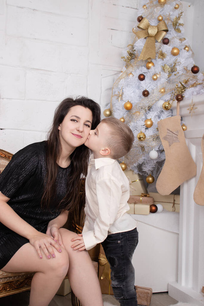 Atmosphere of coming holidays. portrait of happy single mother with her little pleasant boy in white bright room near christmas tree in golden tones - 写真・画像