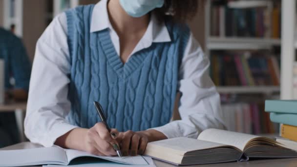 Κοντινό πλάνο αγνώριστη νεαρή γυναίκα σε ιατρική προστατευτική μάσκα κάθεται στο γραφείο στη βιβλιοθήκη άγνωστη φοιτήτρια κορίτσι γράφει αφηρημένη από το βιβλίο στο σημειωματάριο προετοιμασία κατ 'οίκον να σημειώσεις στο βιβλίο εργασίας - Πλάνα, βίντεο