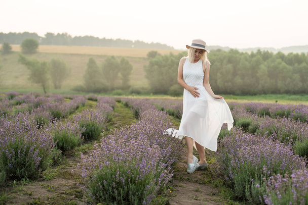 Krásná mladá zdravá žena s bílými šaty radostně běží levandulovým polem, slaměný klobouk - Fotografie, Obrázek