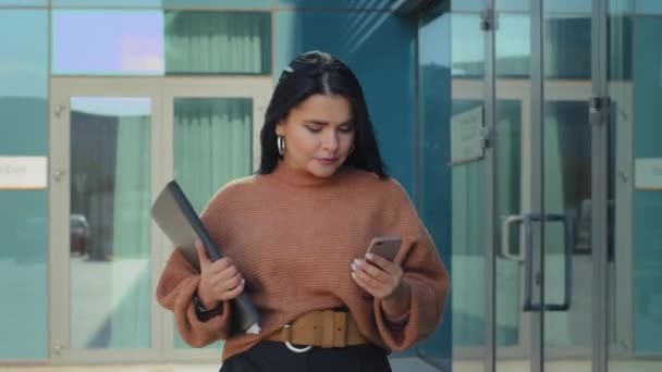 Young confident businesswoman walking down street against background of building carries documents in hands serious focused hispanic girl checking email using mobile phone writes message on smartphone - Imágenes, Vídeo