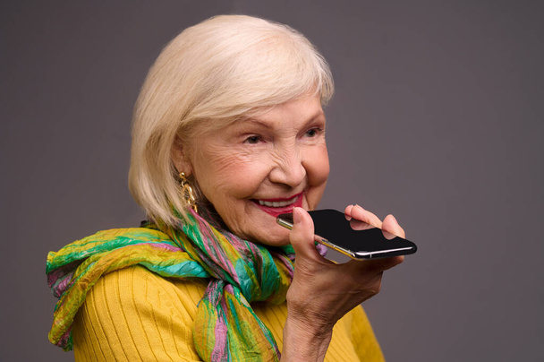 Sarı tişörtlü ve yeşil eşarplı yaşlı kadın sesli mesaj kaydediyor. - Fotoğraf, Görsel