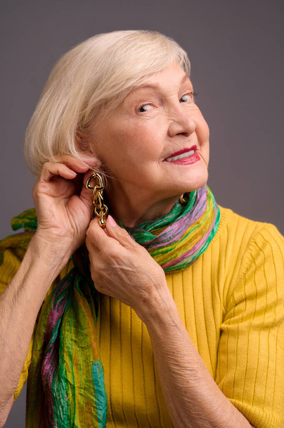 Blone smiling senior woman putting on long earrings - Foto, Imagen