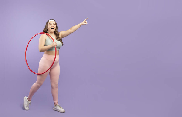 Happy millennial european plus size woman in sports uniform with hula hoop pointing to blank space - Photo, Image