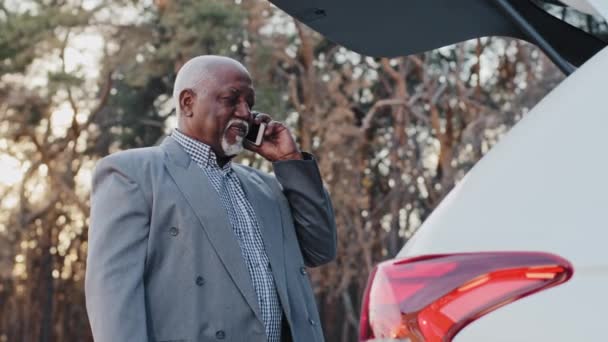 Close-up african american businessman stands outdoors near open trunk of car elderly man entrepreneur talking on mobile telephone business conversation using smartphone mature male answers phone call - Materiaali, video
