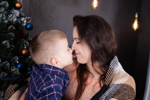 Young mother in a beige knitted sweater near a Christmas tree with her son. single mother with child. family time. mothers love - Photo, Image