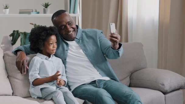 African American family father and daughter sitting in room on couch chatting on webcam mature man with little cute girl shoots video for blog looking at smartphone screen waving hello to phone camera - Filmati, video