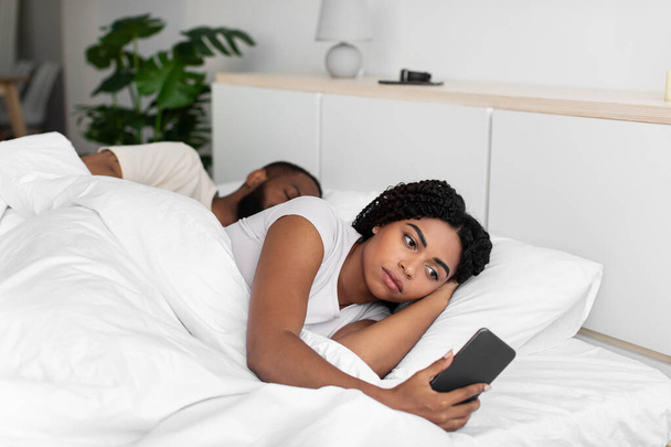 Sad young african american lady looks at smartphone lie on comfortable bed hide cheating - Foto, Imagen