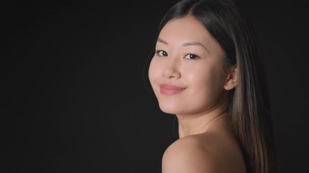 Junge Koreanerin mit perfekter Haut posiert vor schwarzem Hintergrund, lächelt mit nackten Schultern in die Kamera, leerer Raum - Filmmaterial, Video