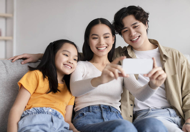Glad smiling young asian couple and teen daughter making photo on phone on sofa in living room - Photo, Image