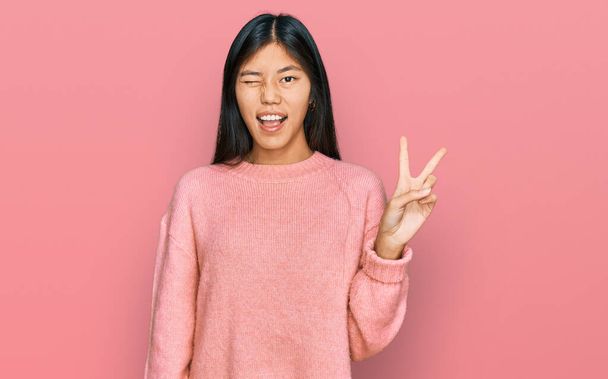 Krásná mladá asijská žena na sobě ležérní zimní svetr s úsměvem šťastný obličej mrkající na kameru dělat vítězství znamení. číslo dvě.  - Fotografie, Obrázek
