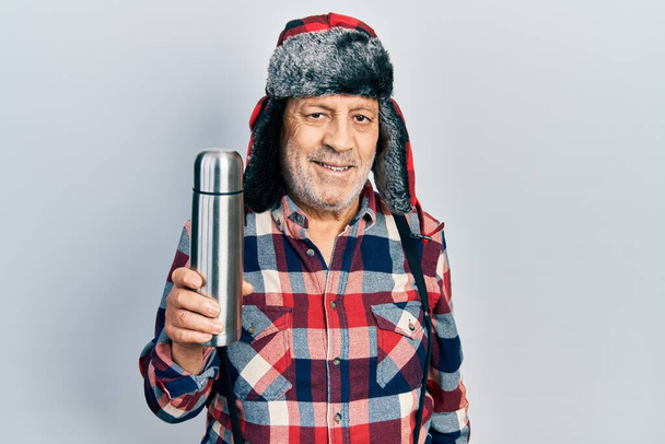 Handsome mature handyman wearing winter hat with ear flaps holding thermo looking positive and happy standing and smiling with a confident smile showing teeth  - Foto, immagini