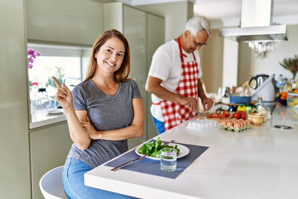 Orta yaşlı İspanyol çift evde sağlıklı salata yiyor. Gülümsüyor, mutlu bir şekilde işaret ediyor.  - Fotoğraf, Görsel