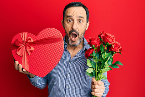 Middle age hispanic man holding valentine day gift and flowers afraid and shocked with surprise and amazed expression, fear and excited face.  - 写真・画像