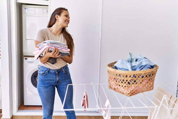 Young hispanic girl doing laundry holding folded clothes at home. - 写真・画像