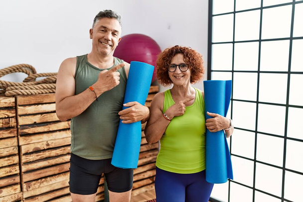 Middle age couple holding yoga mat cheerful with a smile on face pointing with hand and finger up to the side with happy and natural expression  - Foto, Bild