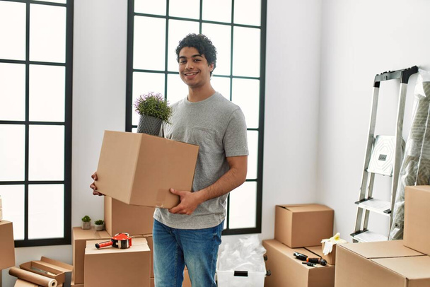 Молодий іспаномовний чоловік посміхається щасливий тримає картонну коробку в новому будинку
. - Фото, зображення