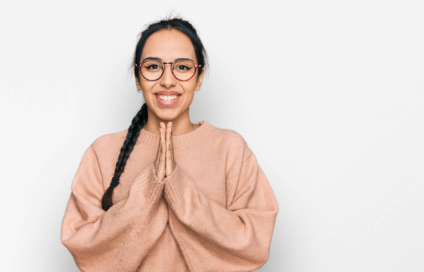 Young hispanic girl wearing casual clothes and glasses praying with hands together asking for forgiveness smiling confident.  - Photo, Image