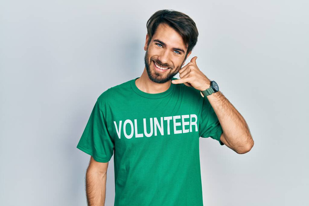 Junger hispanischer Mann im freiwilligen T-Shirt lächelt und macht Telefongesten mit Händen und Fingern, als spräche er am Telefon. Vermittlung von Konzepten.  - Foto, Bild