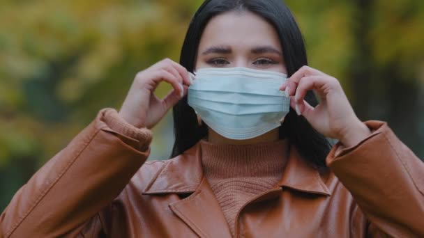 Close-up young woman stands outdoors in park takes off medical protective mask from face throw respirator smiling hispanic girl inhales deeply Fresh air enjoys freedom end quarantine pandemic over - Video, Çekim