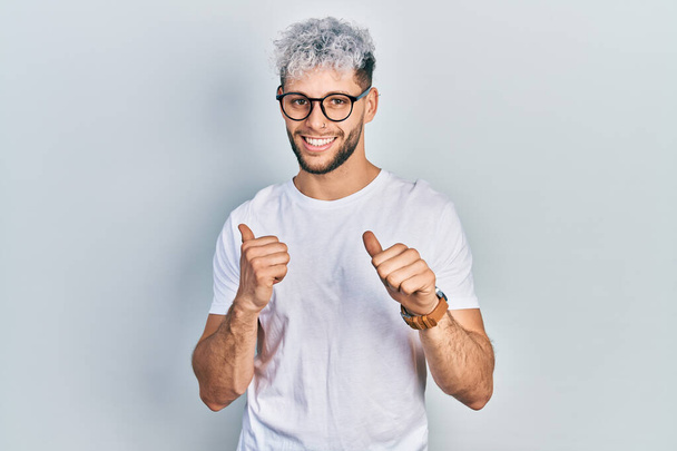 Young hispanic man with modern dyed hair wearing white t shirt and glasses pointing to the back behind with hand and thumbs up, smiling confident  - Fotografie, Obrázek