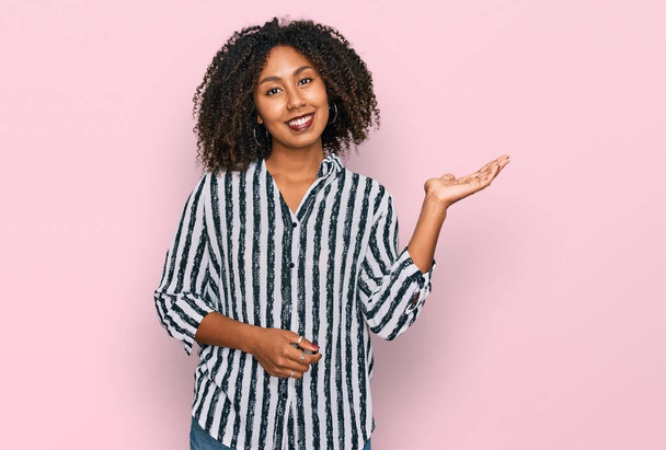 Молодая африканская американка в повседневной одежде улыбается веселым подарком и указывает пальцем на камеру.  - Фото, изображение