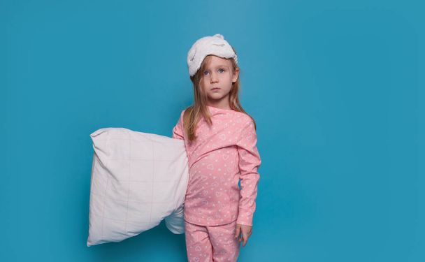 Sad girl in pajamas and with a pillow in her hands stands on a blue background - 写真・画像