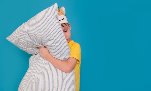 Girl in pajamas and with an eye mask stands on a blue background with a pillow - Fotoğraf, Görsel