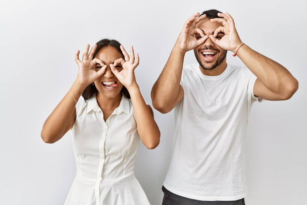 Young interracial couple standing together in love over isolated background doing ok gesture like binoculars sticking tongue out, eyes looking through fingers. crazy expression.  - Foto, Bild