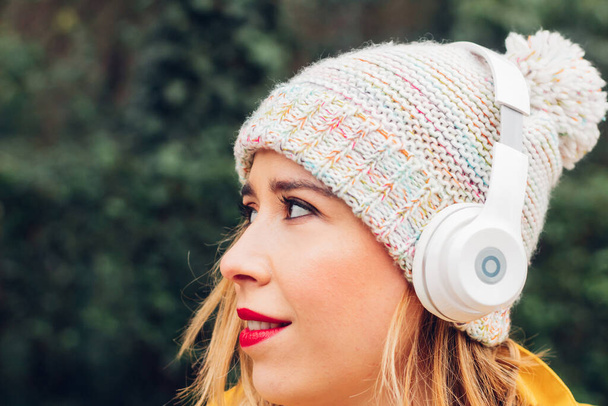 Tvář portrét moderní mladé dospělé ženy na sobě zimní klobouk a bezdrátové sluchátka hledá kopírovat prostor s uvolněným úsměvem tvář venku. - Fotografie, Obrázek