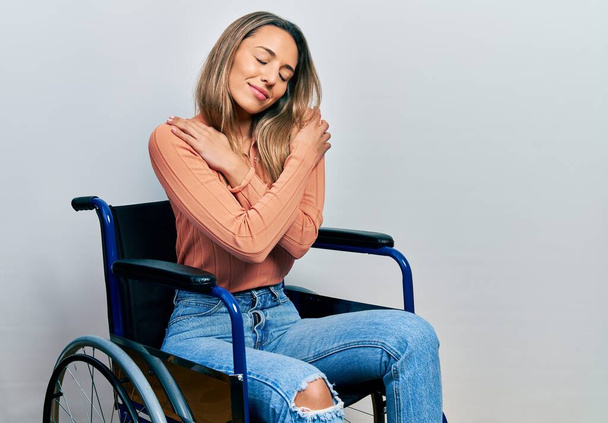 Όμορφη Ισπανίδα που κάθεται σε αναπηρική καρέκλα αγκαλιάζοντας τον εαυτό της χαρούμενο και θετικό, χαμογελώντας με αυτοπεποίθηση. αυτοαγάπη και αυτοφροντίδα  - Φωτογραφία, εικόνα