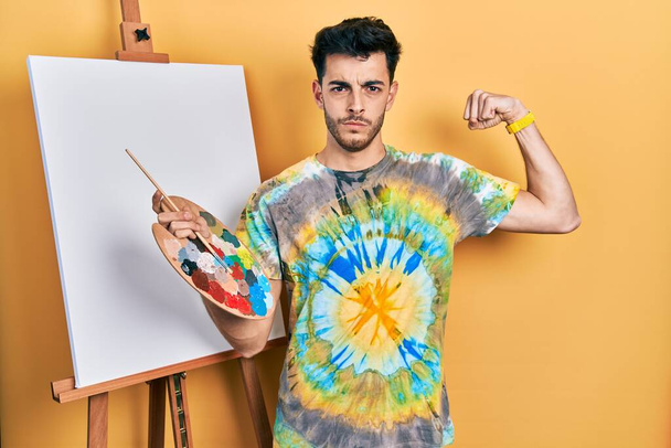 Young hispanic man standing drawing with palette by painter easel stand strong person showing arm muscle, confident and proud of power  - Photo, Image