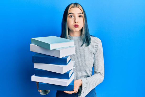 Young modern girl holding a pile of books puffing cheeks with funny face. mouth inflated with air, catching air.  - Foto, Imagen