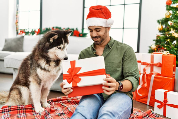 Junger hispanischer Mann packt Geschenk aus und sitzt zu Hause mit Hund am Weihnachtsbaum - Foto, Bild