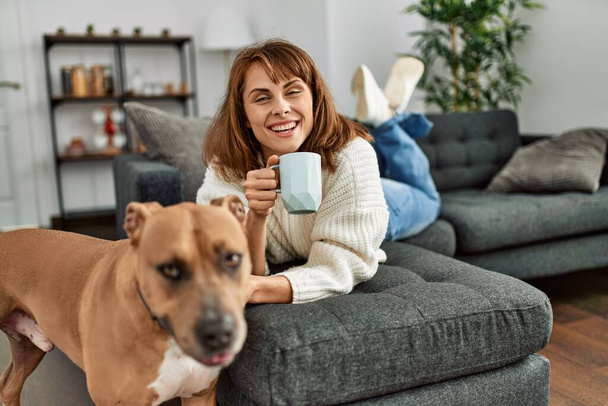 Jonge blanke vrouw drinken koffie liggend op de bank met hond thuis - Foto, afbeelding