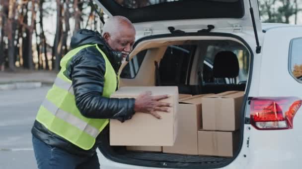 Close-up mature delivery service worker unloading cardboard boxes from trunk of car elderly african american courier loader delivering goods handyman smiling in work clothes accepts shipping outdoors - 映像、動画