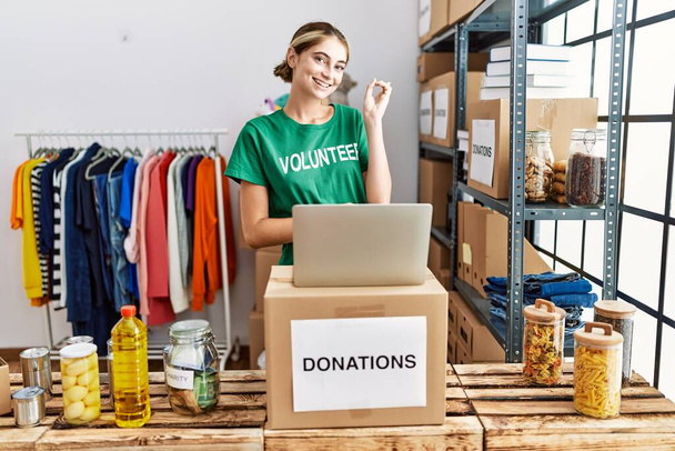 Young blonde woman wearing volunteer t shirt at donations stand smiling happy pointing with hand and finger to the side  - Fotografie, Obrázek