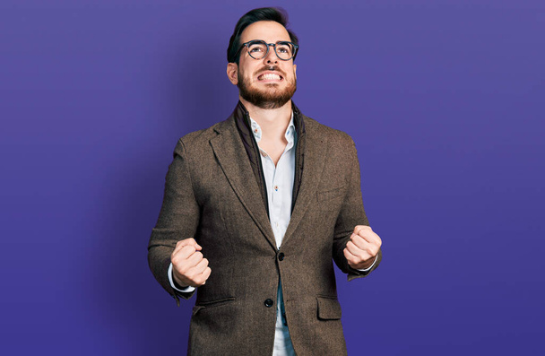 Молодой латиноамериканец в деловой куртке и очках очень счастлив и взволнован, делая жест победителя с поднятыми руками, улыбаясь и крича об успехе. празднование концепции.  - Фото, изображение