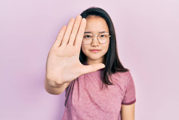 Young chinese girl wearing casual clothes and glasses doing stop sing with palm of the hand. warning expression with negative and serious gesture on the face.  - Фото, изображение
