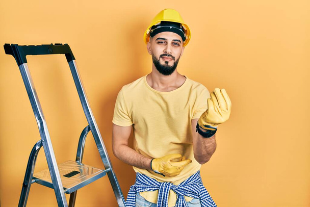 Handsome man with beard by construction stairs wearing hardhat doing italian gesture with hand and fingers confident expression  - Foto, imagen