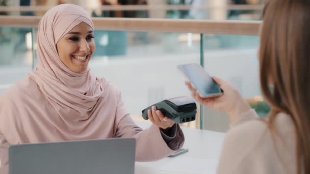 Friendly muslim woman in hijab seller agent offers pay for service through bank terminal girl shopper consumer pays for purchase using contactless payment technology on smartphone client uses phone - Séquence, vidéo