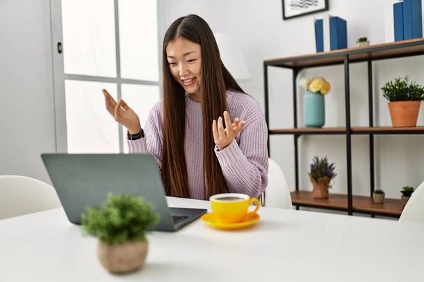 Nuori kiinalainen tyttö, jolla on videopuhelu kannettavan tietokoneen avulla istuu pöydällä kotona. - Valokuva, kuva