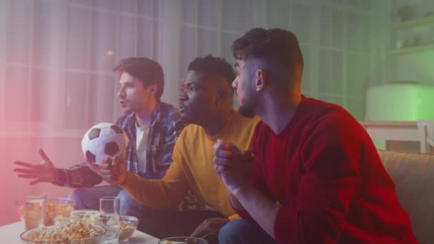 Emotional diverse young men watching TV with sport at night and drinking beer, supporting favorite team with soccer ball - Video