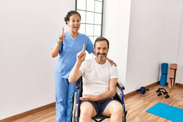 Hispanic middle age man sitting on wheelchair and nurse at rehabilitation clinic surprised with an idea or question pointing finger with happy face, number one  - Photo, Image