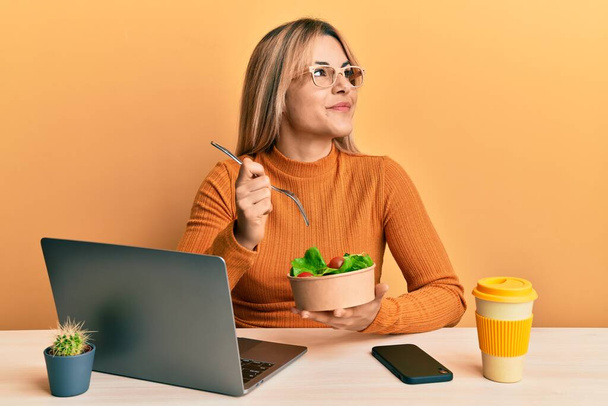 Молода біла жінка, працюючи в офісі, їсть здоровий салат, усміхаючись дивиться в бік і дивиться на мислення
.  - Фото, зображення