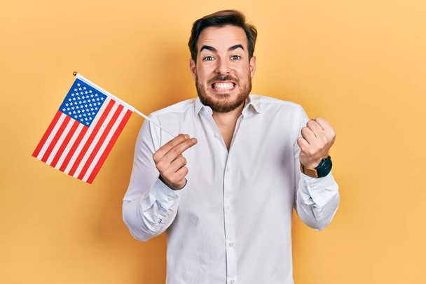 Schöner kaukasischer Mann mit Bart, der stolz die Flagge der Vereinigten Staaten hochhält und mit erhobenem Arm Sieg und Erfolg feiert  - Foto, Bild