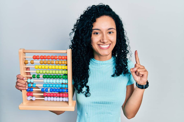 Mulher hispânica jovem com cabelo encaracolado segurando abacus tradicional sorrindo com uma ideia ou pergunta apontando dedo com rosto feliz, número um  - Foto, Imagem