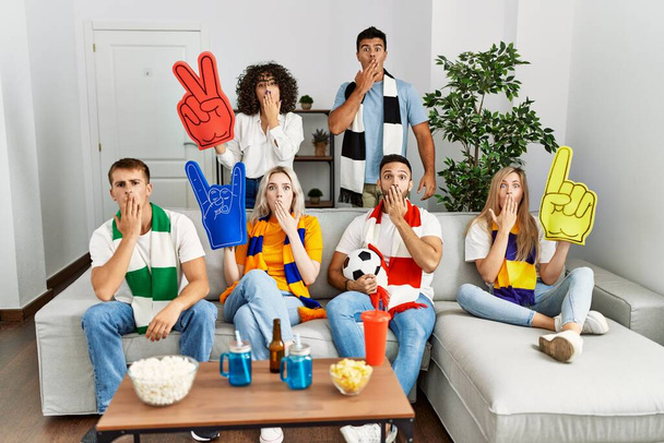 Ομάδα φίλων που υποστηρίζουν την ομάδα ποδοσφαίρου βλέποντας τηλεόραση al σπίτι κάθεται στον καναπέ που καλύπτει το στόμα με το χέρι, σοκαρισμένος και φοβισμένος για λάθος. έκπληκτη έκφραση  - Φωτογραφία, εικόνα