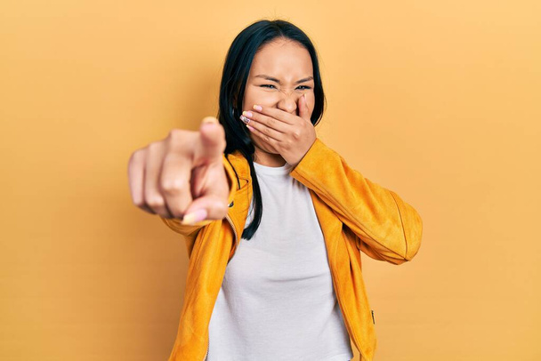 Beautiful hispanic woman with nose piercing wearing yellow leather jacket laughing at you, pointing finger to the camera with hand over mouth, shame expression  - Photo, Image