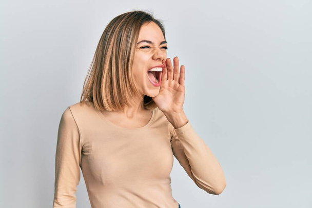 Junge kaukasische blonde Frau in lässigem Pullover schreit und schreit laut Seite an Seite mit der Hand auf dem Mund. Kommunikationskonzept.  - Foto, Bild