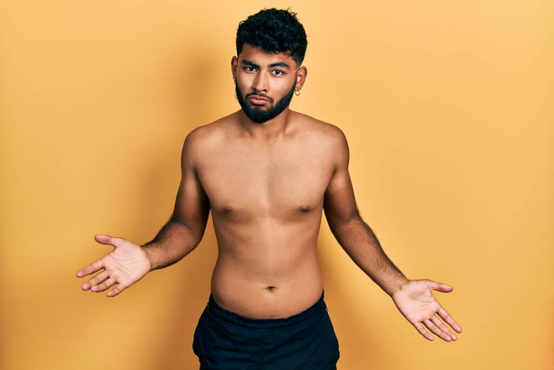 Arab man with beard wearing swimwear shirtless clueless and confused expression with arms and hands raised. doubt concept.  - Foto, immagini